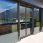 Automatic sliding glass door with a sleek black metal frame, providing access to a modern industrial or commercial space. The reflective glass panels showcase a scenic mountain landscape, enhancing the aesthetic appeal.