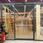 Automatic sliding glass door entrance to a wine section in a supermarket, featuring a modern black frame and transparent panels for clear visibility of wooden shelves stocked with wine bottles.